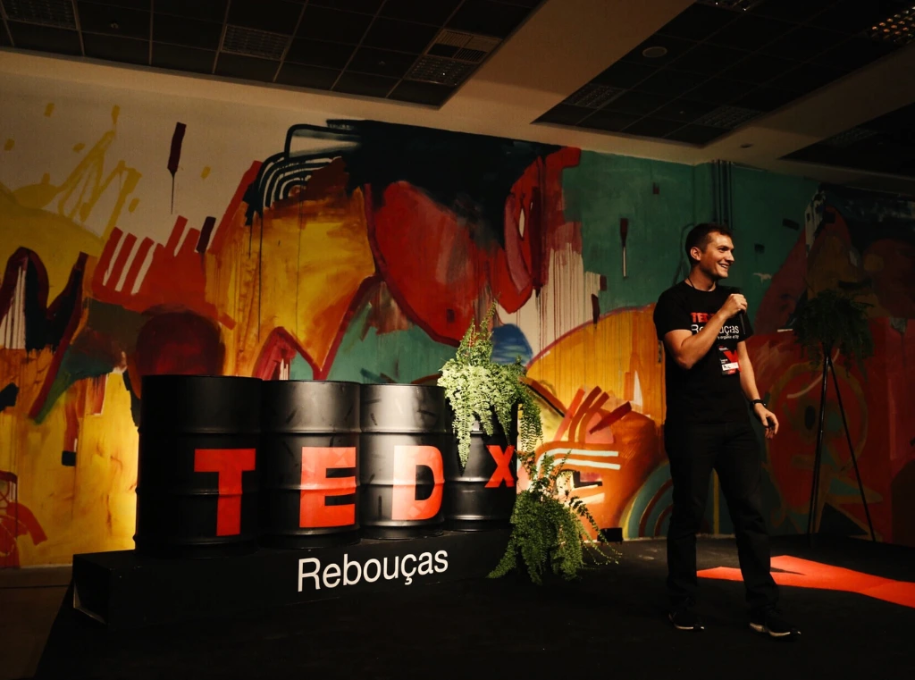 Na Aldeia Coworking Curitiba, apresentador no palco do evento TEDx Rebouças com mural artístico ao fundo.