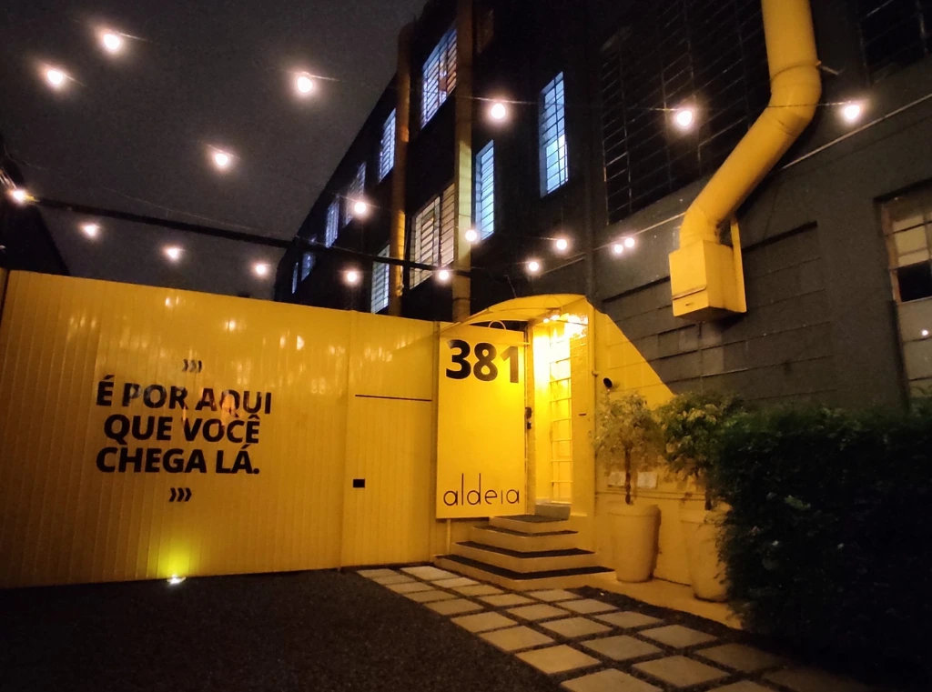 Entrada da Aldeia Coworking em Curitiba com fachada iluminada e frase motivacional 'É por aqui que você chega lá'.