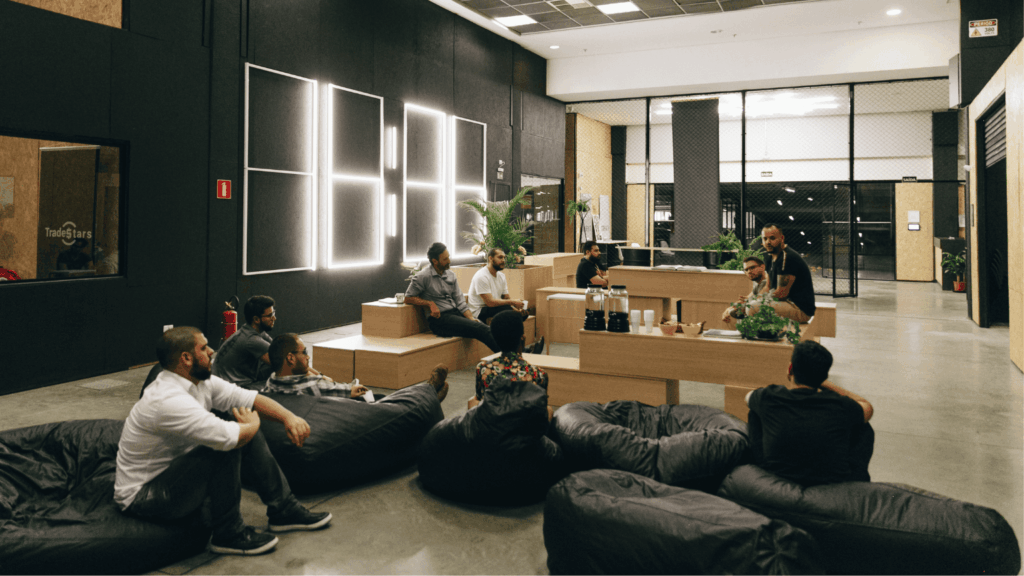 Pessoas reunidas em uma área comum da Aldeia Coworking Estação em Curitiba, sentadas em pufes e bancos de madeira, em um ambiente moderno e colaborativo com iluminação criativa e plantas decorativas.