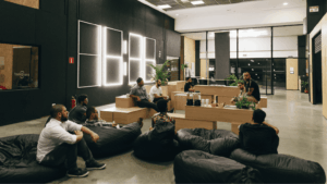 Pessoas reunidas em uma área comum da Aldeia Coworking Estação em Curitiba, sentadas em pufes e bancos de madeira, em um ambiente moderno e colaborativo com iluminação criativa e plantas decorativas.